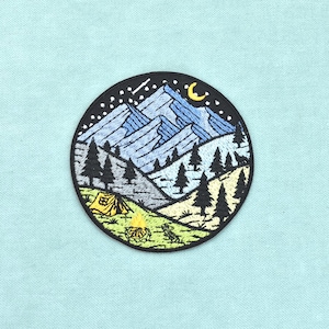 Puede incluir: Un parche bordado circular que presenta un paisaje de montaña nocturno. El diseño incluye montañas azules, pinos negros, una tienda de campaña amarilla y una fogata bajo un cielo negro con estrellas blancas y una luna creciente amarilla, bordeado en negro.