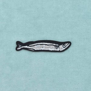 Op de afbeelding: Een geborduurde patch met een kleine, slanke vis met een zwarte omlijning en details van zilvergrijs garen. De vis is horizontaal afgebeeld en toont een eenvoudig oog, een gedefinieerde vin en een gesegmenteerde staartstructuur.