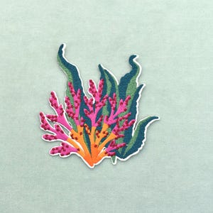 May include: Embroidered patch featuring a colorful coral reef design. The patch showcases vibrant pink, orange, and green hues, with a white border. The design includes detailed coral branches and seaweed-like elements, perfect for adding to clothing or accessories.