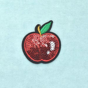Op de afbeelding: Een rode appelpatch met glanzende lovertjes en een groen geborduurd blaadje. De appelvorm is omlijnd met zwart garen, wat een duidelijke rand creëert. Het oppervlak van de vrucht is bedekt met reflecterende rode lovertjes met witte accenten.