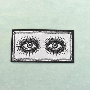 Puede incluir: Parche bordado con dos ojos estilizados con largas pestañas y líneas radiantes. Los ojos son negros sobre un fondo blanco, enmarcados por un borde negro. El parche rectangular es adecuado para ropa o accesorios.