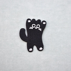 Peut inclure: Écusson de chat noir avec des détails brodés blancs. Le chat est debout, les pattes en l'air et la queue courbée. Le visage du chat a des moustaches et des yeux blancs. L'écusson est sur un fond gris clair. Parfait pour les vêtements ou accessoires.
