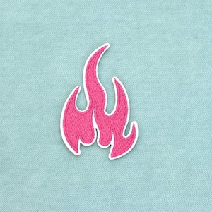 Peut inclure: Un écusson brodé rose vif en forme de flamme stylisée. Le motif de flamme présente un sommet pointu et des bords incurvés et dentelés, souligné par une fine bordure blanche qui définit la forme sur le tissu bleu clair environnant.