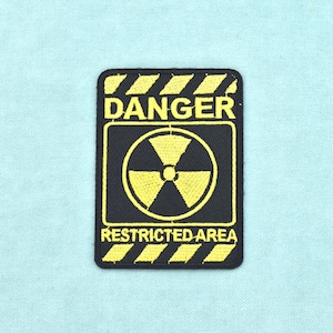 Puede incluir: Un parche bordado rectangular con fondo negro, bordes y texto en amarillo. Las secciones superior e inferior tienen rayas diagonales amarillas. El texto dice DANGER y RESTRICTED AREA junto a un símbolo de radiación amarillo central.