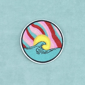 Peut inclure: Un écusson brodé circulaire représentant une vague océanique stylisée en bleu clair, un soleil jaune vif et un ciel rempli de bandes ondulées de fil rose, rouge et bleu clair. Le motif est entouré d'une bordure blanche épaisse.