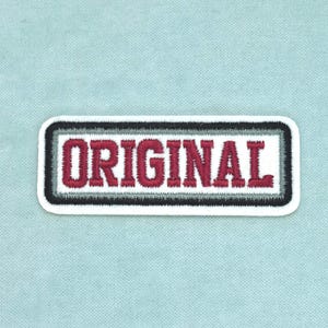 Peut inclure: Un écusson rectangulaire brodé portant le mot ORIGINAL en lettres capitales rouge foncé. Le texte est centré sur un fond blanc, entouré d'une fine bordure grise et d'une bordure extérieure noire plus épaisse aux coins arrondis.