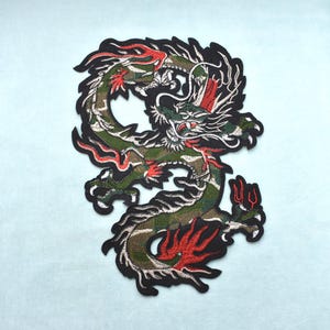 Op de afbeelding: Geborduurde drakenpatch in groen, rood en zwart. Het drakenontwerp heeft ingewikkelde details en een camouflagepatroon op het lichaam. De patch heeft een zwarte rand en is geplaatst tegen een lichtblauwe achtergrond.