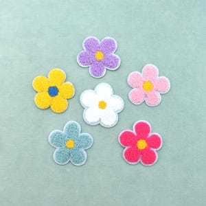 May include: A set of six flower-shaped patches with a textured, fuzzy surface. The patches feature various colours including purple, yellow, pink, white, light blue, and hot pink, each with a contrasting centre colour and a white border edge.