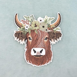 May include: Embroidered patch of a Highland cow's head, wearing a floral crown. The cow has brown fur, large horns, and a white face. The floral crown features green, white, and brown flowers and foliage. The patch is set against a light blue backdrop.
