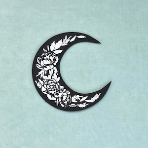 Peut inclure: Un écusson en forme de croissant de lune avec un fond noir et un motif floral brodé en blanc. Le motif se compose de fleurs ressemblant à des pivoines et de feuilles détaillées disposées le long de la courbe de la lune aux bords nets.