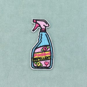 Peut inclure: Un écusson brodé en forme de flacon pulvérisateur avec un corps bleu clair, un embout rose et une étiquette rose. L'étiquette porte le texte "flickboy REPELLENT" en noir, agrémenté de petites fleurs brodées et d'une icône de cœur rose.