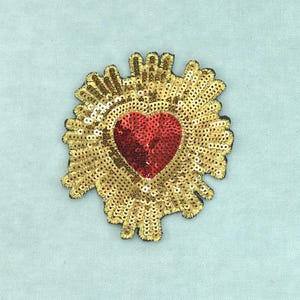 Può includere: Una toppa a forma di cuore ricoperta di paillettes rosse, circondata da un design a raggiera realizzato con paillettes dorate. L'intero pezzo presenta una texture densa e scintillante con un bordo definito, creando un emblema decorativo.