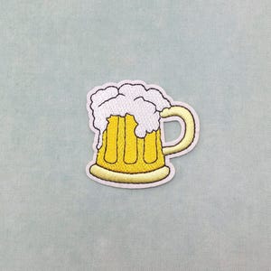May include: Embroidered patch depicting a yellow beer mug with white foam spilling over the top. The mug has a curved handle and is outlined in a dark yellow. The patch is set against a light blue backdrop.