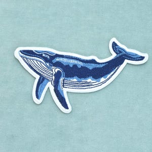 Könnte beinhalten: Ein gestickter Aufnäher, der einen Buckelwal in verschiedenen Blau-, Marine- und Hellblautönen zeigt. Der Wal ist in einer Schwimmposition mit ausgestreckten Flossen und sichtbarer Schwanzflosse dargestellt, umrandet von einem weißen Rand auf hellblauem Grund.