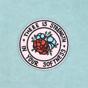 Könnte beinhalten: Ein kreisförmiger bestickter Aufnäher mit hellrosa Hintergrund und schwarzem Rand. In der Mitte befindet sich eine rote und blaue Blume mit grünen Blättern. Der Text "THERE IS STRENGTH IN YOUR SOFTNESS" ist in schwarzen Großbuchstaben am Rand gestickt.