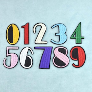 Può includere: Un set di dieci toppe ricamate con i numeri da zero a nove, ognuna con una combinazione di colori distinta che include giallo, rosso, crema, azzurro, verde, rosa, bianco, viola e nero, disposte su due file.