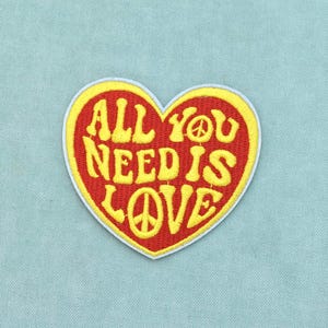 Può includere: Una toppa ricamata a forma di cuore con sfondo rosso e bordo giallo. Il testo ALL YOU NEED IS LOVE è ricamato in giallo, con un simbolo di pace che sostituisce la lettera O nelle parole YOU e LOVE, rifinito con un bordo bianco.