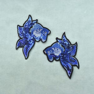 Op de afbeelding: Twee geborduurde vispatches met levendige blauwe pailletten en draaddetails. Elke patch heeft de vorm van een kempvis met vloeiende vinnen en een zwarte rand, met gedetailleerde schubpatronen en een opvallend oog van witte draad.