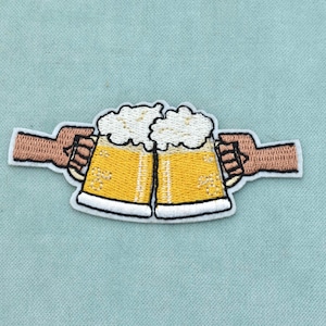 May include: An embroidered patch featuring two yellow beer mugs with white foam being held by two hands in a cheers gesture. The design has a white border and is stitched with yellow, white, brown, and black thread on a light blue fabric background.