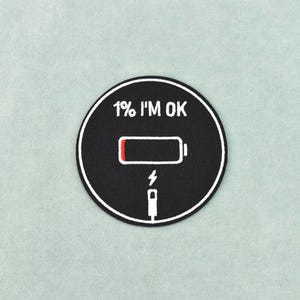 Puede incluir: Un parche circular negro con borde blanco, con el texto "1% I'M OK" sobre un icono de batería baja con relleno rojo. Debajo de la batería hay un rayo y un icono de puerto de carga.