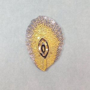 May include: A decorative, leaf-shaped embellishment with a grey base and intricate gold embroidery. The design features a central oval with black and gold accents, and is adorned with small, gold sequins.
