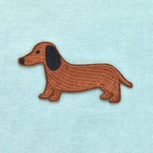 Peut inclure: Un écusson brodé thermocollant en forme de chien teckel. Le chien possède un corps marron avec une oreille noire et un petit nez noir. La broderie présente des points denses et texturés sur toute la forme de la silhouette canine.
