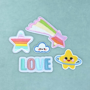 Op de afbeelding: Een verzameling geborduurde patches met een regenboogkleurige ster, een vallende ster met een pastelkleurige staart, een kleine lachende wolk, het woord LOVE in pastelkleurige letters en een gele ster met een blauwe ring en gezicht.