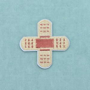 Op de afbeelding: Een geborduurde patch in de vorm van een kruis, bestaande uit twee elkaar kruisende pleisters. De patch heeft een beige basis met een donkerder roodbruin vierkant in het midden en kleine rode stikdetails op de uiteinden van elke arm.