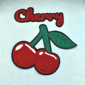 Puede incluir: Un parche de chenilla rojo con la palabra Cherry en letra cursiva, colocado sobre un par de cerezas rojas con tallos verdes y una hoja verde, todo delineado en negro con reflejos blancos en la superficie de las frutas.