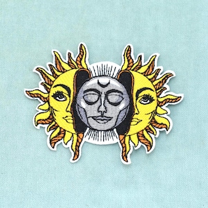 Puede incluir: Un parche bordado que presenta una cara de luna gris central con un símbolo de media luna en la frente, flanqueada por dos caras de sol amarillas con rayos hacia afuera. El diseño es simétrico con costuras negras detalladas sobre fondo blanco.