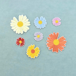 Könnte beinhalten: Eine Sammlung von sieben bestickten Blumenaufnähern in verschiedenen Größen und Farben, darunter Weiß, Hellblau, Rosa, Rot, Violett, Gelb und Orange, jeweils mit einem kontrastierenden runden Zentrum und einem weißen Stickrand.