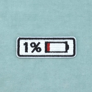 Può includere: Una toppa ricamata rettangolare che presenta un'icona di batteria bianca con un bordo nero. Il design include il testo 1% in nero, con un piccolo segmento rosso all'inizio della barra della batteria, indicando un basso livello di carica.