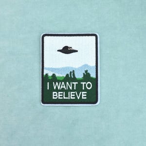 Könnte beinhalten: Ein rechteckiger bestickter Aufnäher mit einer Landschaftsszene, die ein schwarzes UFO zeigt, das in einem hellblauen Himmel über grünen Hügeln und Bäumen schwebt. Der Text I WANT TO BELIEVE ist in weißen Großbuchstaben im grünen Vordergrund.