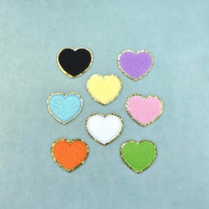 Peut inclure: Huit écussons en forme de cœur disposés en groupe. Chaque écusson présente une surface texturée et duveteuse dans une couleur unie distincte : noir, violet, jaune, bleu clair, rose, blanc, orange et vert. Chaque cœur est bordé de paillettes dorées.
