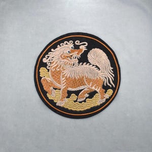 Könnte beinhalten: Gestickter Aufnäher mit einem Fabelwesen, möglicherweise einem Qilin, in Orange-, Beige- und Cremetönen. Das Design befindet sich auf einem schwarzen, kreisförmigen Hintergrund mit einem dünnen orangefarbenen Rand. Die Kreatur ist im Profil dargestellt.