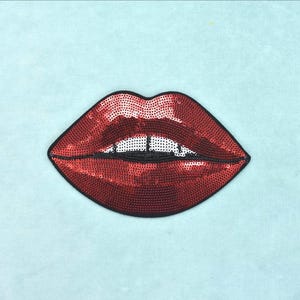 Könnte beinhalten: Ein roter Paillettenaufnäher in Form eines Lippenpaares mit schwarzem Rand. Die Lippen zeigen in der Mitte einen kleinen Bereich aus weißen Pailletten, die Zähne darstellen. Der Aufnäher besteht aus dichten, glänzenden roten Pailletten.