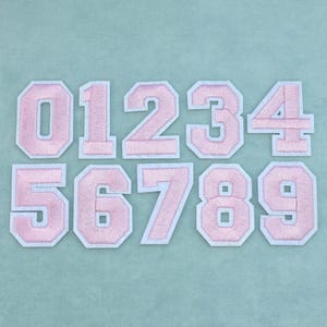 Op de afbeelding: Een set van tien opstrijkbare patches met de cijfers nul tot en met negen in een varsity-stijl lettertype. Elke patch is lichtroze met een witte rand, gerangschikt in twee rijen van vijf op een effen lichtblauwe achtergrond.