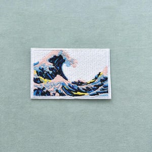 Peut inclure: Écusson brodé représentant une vague stylisée dans des tons de bleu, rose et jaune sur un fond rectangulaire blanc. L'écusson est posé sur un tissu vert clair.