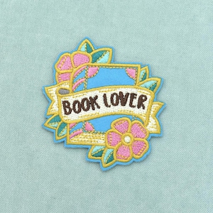 Può includere: Una toppa ricamata raffigurante un libro blu circondato da fiori rosa e foglie verdi. Un nastro color crema si sovrappone al centro del libro con il testo BOOK LOVER ricamato in filo marrone scuro, il tutto contornato da un bordo blu.