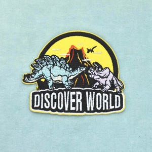 Könnte beinhalten: Ein kreisförmiger bestickter Aufnäher mit einem hellblauen Stegosaurus und einem grauen Triceratops vor einem roten Vulkan unter gelbem Himmel. Auf der schwarzen Basis des Aufnähers steht der weiße Text DISCOVER WORLD in fetter serifenloser Schrift.