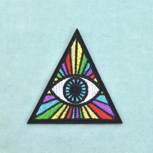 Könnte beinhalten: Ein dreieckiger bestickter Aufnäher mit einem zentralen Auge, das eine schwarze Pupille und eine blaue Iris aufweist. Das Auge ist von einem lebendigen, mehrfarbigen Strahlenmuster in Rot, Gelb, Grün, Blau und Violett umgeben, eingefasst von einem schwarzen Rand.