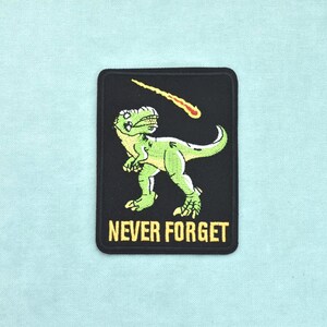 Könnte beinhalten: Ein rechteckiger, bestickter Aufnäher mit schwarzem Hintergrund, der einen grünen Tyrannosaurus Rex zeigt, welcher zu einem gelb-orangen, brennenden Meteor am Himmel aufblickt. Der Text NEVER FORGET ist unten in gelben Großbuchstaben eingestickt.
