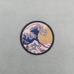 Könnte beinhalten: Ein runder gestickter Aufnäher mit einer weißen Welle mit blauen Akzenten auf einem gelben und rosafarbenen Hintergrund. Die Welle erinnert an die Große Welle vor Kanagawa, ein berühmtes japanisches Holzschnittbild.