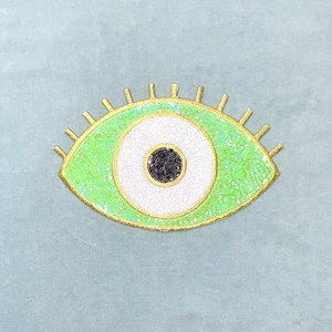 Pode incluir: Um emblema em forma de olho com uma íris de lantejoulas verde brilhante, uma esclera de contas brancas e uma pupila de contas pretas. O design é contornado com fio dourado e inclui cílios dourados na borda superior do olho.