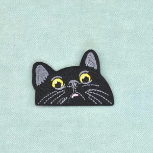 Pode incluir: Um emblema bordado com a cabeça de um gato preto com olhos amarelos grandes, interior das orelhas cinzento, um pequeno nariz cor-de-rosa e bigodes brancos. O gato é representado do peito para cima com uma margem inferior plana.
