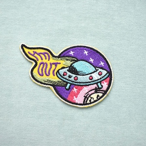 Peut inclure: Écusson brodé représentant un OVNI turquoise avec des lumières rouges, sur fond d'une scène spatiale violette et rose avec des étoiles. Le patch comprend le texte "I'M OUT" en jaune, souligné de noir.