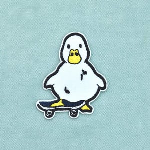 Op de afbeelding: Een geborduurde patch met een witte eend met een gele snavel die op een donkerblauw skateboard staat. De eend heeft zwarte omlijningen en kleine zwarte markeringen op het lichaam. Het ontwerp staat op een effen, lichtblauwe textuurachtergrond.