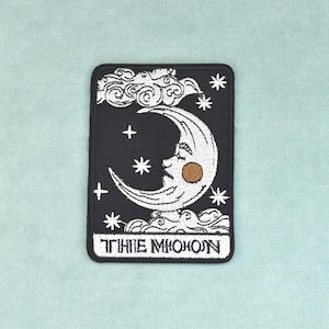 Op de afbeelding: Een rechthoekige geborduurde patch met een zwarte achtergrond met een witte maansikkel met een gezicht en een beige wang. Het ontwerp bevat witte sterren en wolken, met de tekst THE MOON in het wit onderaan de patch.