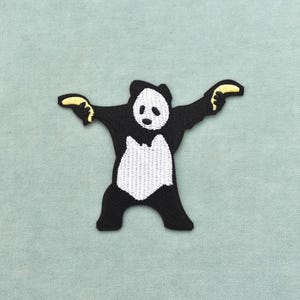 Peut inclure: Un écusson brodé représentant un panda noir et blanc debout, les bras écartés. Le panda tient une banane jaune dans chaque main. Le motif est réalisé avec un travail de fil détaillé sur un fond vert clair uni.