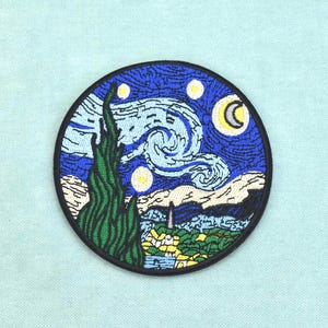Puede incluir: Un parche bordado circular que presenta una reproducción de La noche estrellada. El diseño incluye un alto ciprés verde oscuro en primer plano, un cielo nocturno azul con remolinos, estrellas amarillas, una luna creciente y un pequeño pueblo.